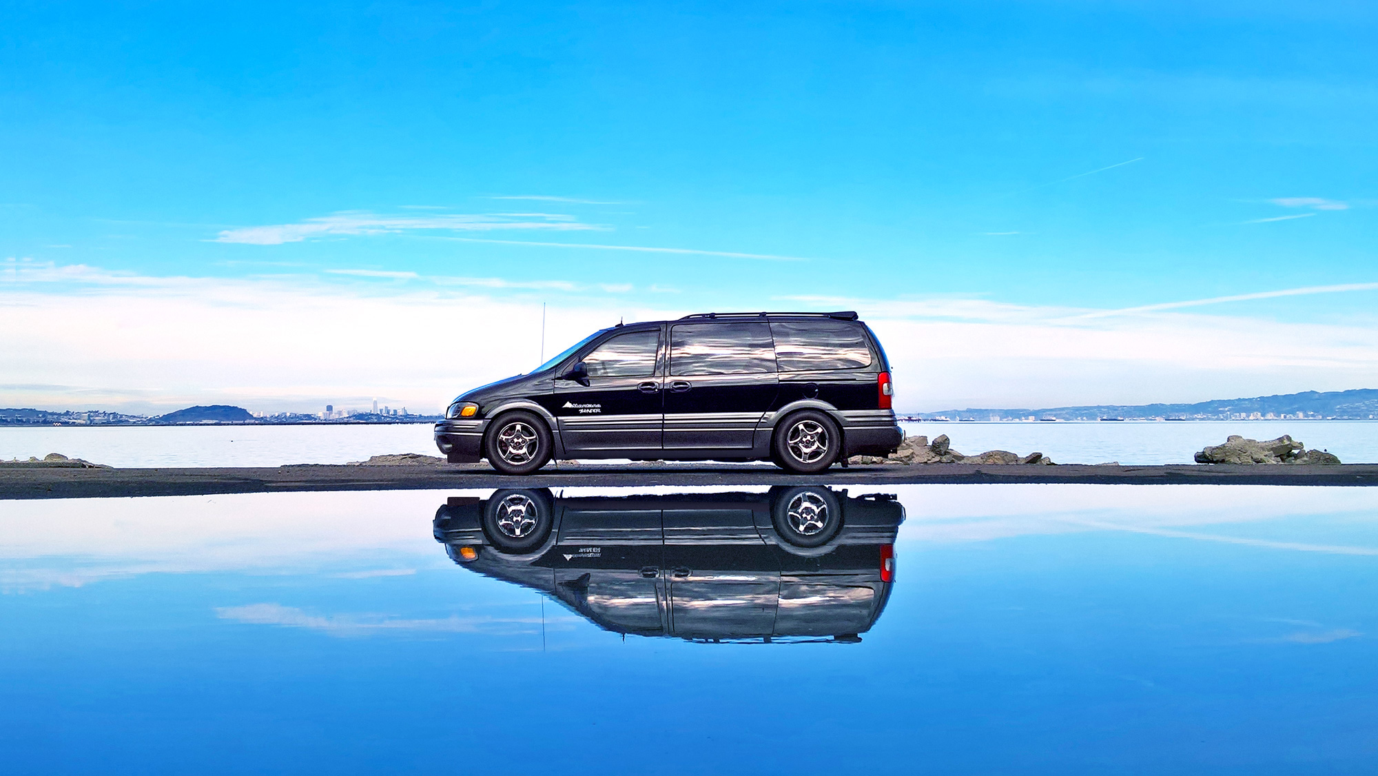 Reflection in water of a profile/side view of a 2003 AWD Pontiac Montana Thunder in black overlooking SFO and San Francisco skyline